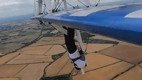 A woman is strapped to the wing of a plane which is flying upside down over brown fields.