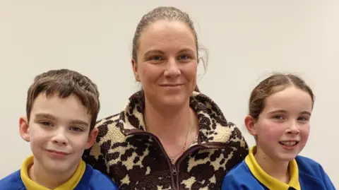 Woman with brown and cream fleece, smiling at camera, two primary school aged children sit either side of her also smiling at camera. Boy left and girl right. Both wearing a blue jumper with a yellow shirt collar. 