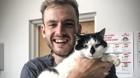 RSPCA Vet Rory Cowlam who is smiling while holding a black and white cat