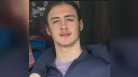 Kailem is smiling and looking at the camera. He has brown hair and is wearing a dark blue jacket and a pink and grey top.