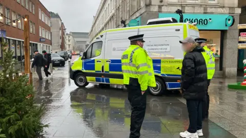 A police van parked on a high street. It says "Facial Recognition in use" on the side. There are two police officers wearing high-vis outside the van talking to a man in a black coat who is wearing a cap. It is raining.