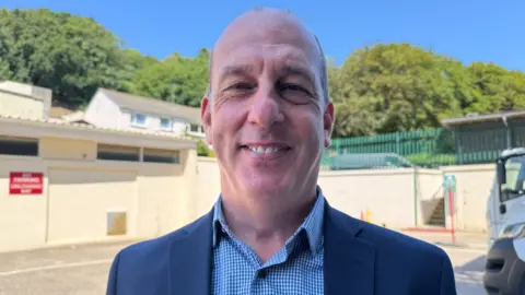 A man standing in front of a white building, wearing a dark blazer and checkered shirt. Trees, a parked vehicle, and a clear blue sky are in the background.