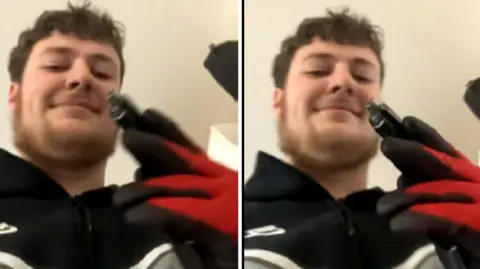 South Wales Police/CPS Huntley has dark brown hair, a beard and is smiling at the camera. He's holding the pistol up the camera and wearing black and red gloves