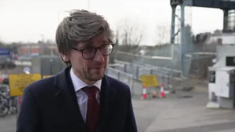 Marcus Jones giving an interview at the Oxford Railway Station. He is wearing a suit with a red tie and glasses. Signs about the closure and diversions can be seen behind him. 