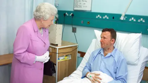 Contributed Mr Lait in a hospital bed as he speaks with Queen Elizabeth II. He is propped up on white pillows and is wearing blue pyjamas. His face is scarred. The Queen is wearing a pink dress and, as she stands by the bed, she is leaning forward slightly as she speaks to Mr Lait.