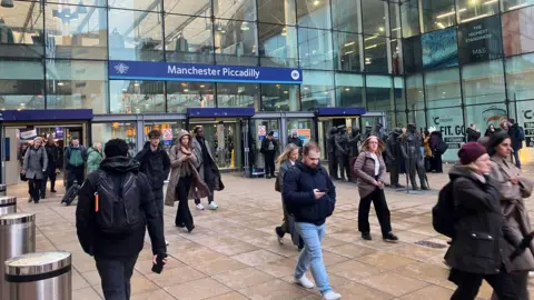 Photograph of commuters at the entrance to Manchester Piccadilly Station. The image shows people moving in and out of the main entrance.
