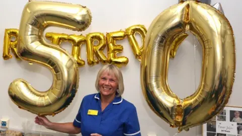Julie is wearing her uniform and is stood in front of balloon letters spelling out that she has retired as she holds the numbers 5 and 0  in front of cakes and various treats.