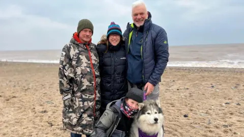 From left, Ely's dad Steven, who is wearing a camouflage coat and a dark green woolly hat. Next to him is mum Vicki, who is wearing a long black coat and bright blue and pink bobble hat. She is stood next to Adrian, who is wearing a navy jacket, and dark grey trousers. All three are smiling and have their arms around each other. In front of them, at their feet, is Ely's brother Guy, who has his arm around Thunder. They are on Hornsea beach.