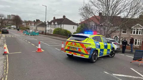 A police car is parked across a road and a police tape can be seen in the distance.