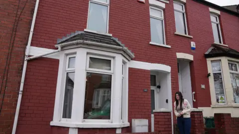 Photo of the outside of a two bedroom terraced house on Trinity Street, Barry, Vale of Glamorgan. The house is red brick with a large bay window at the front, with white window frames and a white front door. Lisa Molloy stands outside the front of the house, holding her hands together in front of her. 