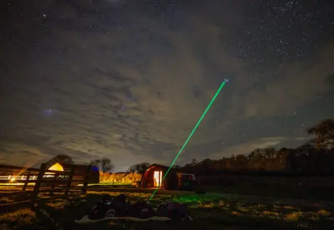 BBC/Thomas Winstone Dafydd Wyn Morgan shines a green laser at the night sky. There are grey clouds obscuring parts of the sky but bright stars are visible. Three people lie on a blanket on the grass. In the background there are huts with lights on visible. 