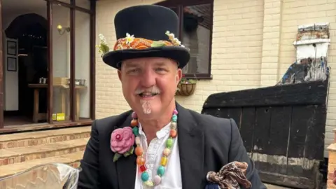 Contributed A man wearing a black top hat, black suit jacket and a white top smiles at the camera. He has some colourful items attached to his jacket including a fake rose, multi-coloured necklace and there is an orange neckerchief tied around the base of his hat.