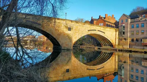 BBC WEATHER WATCHERS/ JACK PROST A large stone bridge over a river has been reflected in the water below. It has been lit in yellow light from the sun and the sky above is a bright blue.