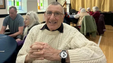 The image shows an older man with glasses wearing a cream cable knit jumper, spectacles and a large watch. He is smiling at the camera 