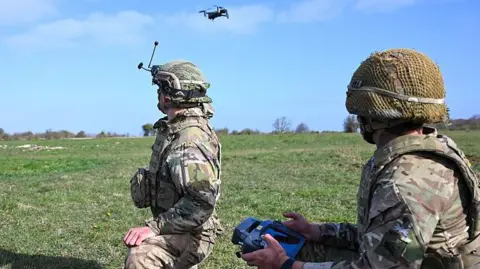 Two soldiers kneel in a field, with one using a remote control to operate a drone.