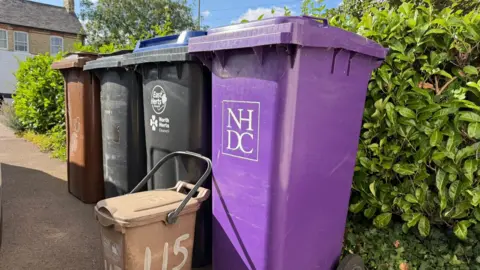 Ben Marsden Four wheelie bins are lined up against a hedge. One is purple, two are black, and one is brown. They are variously branded North Herts District Council and East Herts Council. Standing in front of them is a much smaller brown food waste caddy with a handle and the number "115" hand-painted on it.