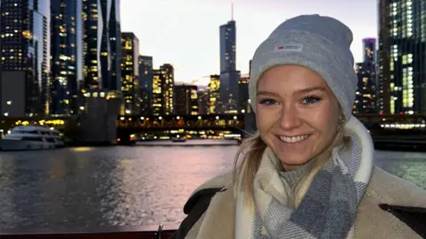 Lily, a young woman, is smiling. She has blonde hair and is wearing a grey woollen hat and scarf. In the background are skyscrapers and a river. 