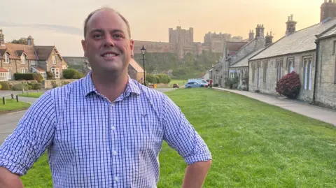 Guy Renner-Thompson is a man in his 30s with a receding hairline and wearing a blue shirt. He is standing in front of the very large Bamburgh castle, on either side are the cottages of the village