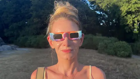 BBC / Phil Harrison Rhiannon Quinche stands in a grassy clearing on Tunbridge Wells Common wearing eclipse glasses, with its famous rocks in the background. Trees surround the area and warm evening sunlight falls across the landscape.