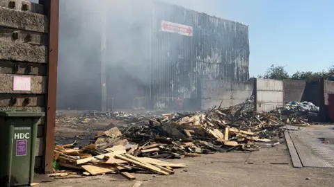 Richard Knights/BBC Piles of charred wood in front of a burnt out corrugated iron building.