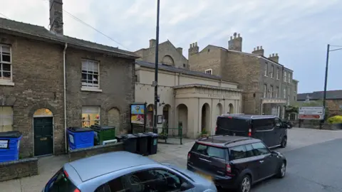 Google A street view of Westgate Street. A small theatre can be seen in a cream colour and has an arched entrance. Homes made of brick can be seen either side of the theatre. Cars are parked outside the theatre and homes. 
