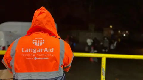 Khalsa Aid A person is wearing an orange hi-viz jacket with silver strips, standing at a yellow balcony rail, The hi-viz jacket reads, "Langar Aid, feeding humanity, a project by Khalsa Aid."
