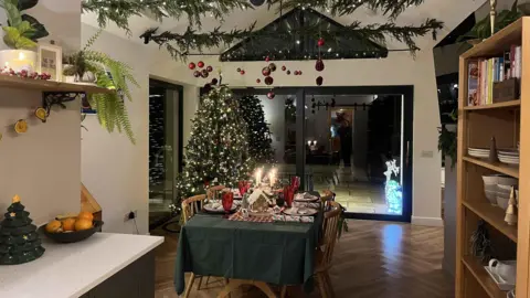 IWC Media A Christmassy kitchen with a vaulted ceiling. Red Christmas baubles dangle from the ceiling, which is also decorated with Christmas foliage. In the corner is a large Christmas tree decorated with hundreds of Christmas lights. A dining table, with a green tablecloth, is garnished with candles and a gingerbread house. 