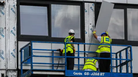 Three workmen in hi-vis jackets and helmets removing cladding from a building