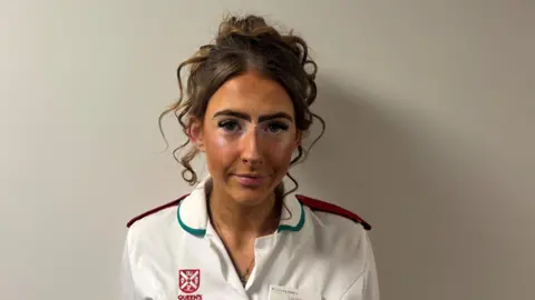 Louise Bradford is wearing a white nurses uniform. She has brown hair. She is in front of a white background. 