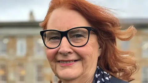 Anne Robson smiles at the camera. She has shoulder length red hair and black-framed glasses. She is wearing a navy suit jacket with navy scarf decorated with white dots. Behind her is a large Georgian-style manor house