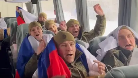 EPA/Shutterstock A handout screenshot from a video released by the Russian defence ministry shows Russian soldiers - draped in Russias national white-blue-and-red flags - riding on a bus after a prisoner exchange with Ukraine. Photo: 5 February 2026