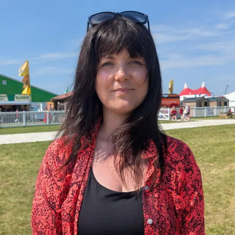 Helen Jenner at the Eisteddfod. She smiles whilst looking at camera and wears a red blouse, black sunglasses on her head and has dark brown hair.