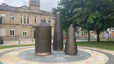 BBC Three large metal sculptures stand in a circular paved area in front of a sandstone building. The three sculpture are a bronze colour, one is a cylinder shape which tapers towards the top. The other two are more conical in shape. 