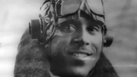 Eddy Smythe Johnny Smythe looks slightly off camera, smiling. It's a black and white image, he's wearing his flying uniform with a helmet and goggles on his forehead and a sheep skinned fluffy collar of his jacket