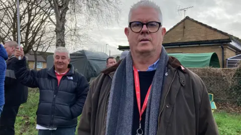Mike Hobbs, who has grey hair, is wearing dark-rimmed glasses, a scarf and an open jacket with a red lanyard. He is looking into the camera and striking staff can be seen in the background.