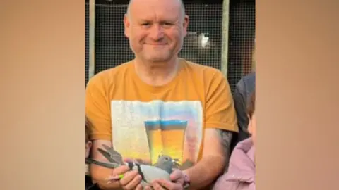 Danny Rodgers Danny Rodgers, a man in his 50s with stubble and a shaved head, smiles and holds a pigeon, which sits in his hands. He is wearing a bright orange shirt with a graphic on the front.