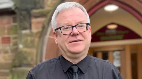 A grey haired man in glasses with a black shirt and tie stands in front of an arched sandstone doorway