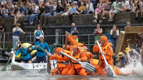 Paul Box Two teams paddling across the river during a raft race. One team is dressed as beans on toast, in bright orange overalls with cutlery oars, and the other's raft looks like a swan. 