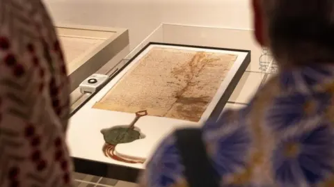 Durham Cathedral A page from the Magna Carta on display at the Durham Cathedral Museum. The page is old and stained with age. A large metal seal has been placed at the bottom of the display. Two people are looking at the exhibition.