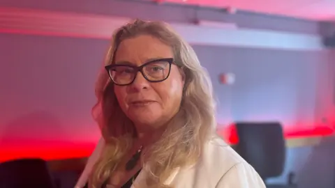 A blonde woman with black rimmed glasses and a cream jacket