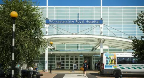 The outside of Worcester royal hospital where a woman in a black dress is walking across a zebra crossing. There is a silver ambulance to the right of the road