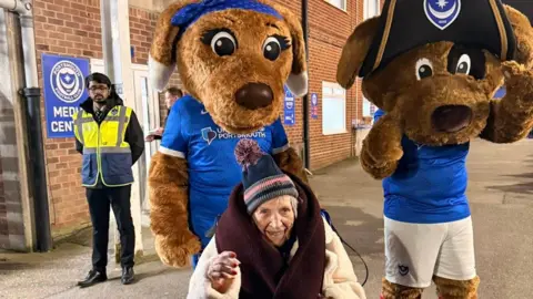Cosham Court Nursing Home Joan is pictured seated outside Portsmouth's stadium. Behind her are two mascots dressed as giant dogs and wearing Pompey strips. 