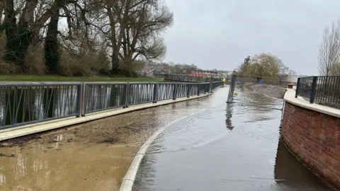 Water from a river is spilling onto a path. There is a barrier in between the river and the path. A man wearing high-vis can be seen walking in the distance.