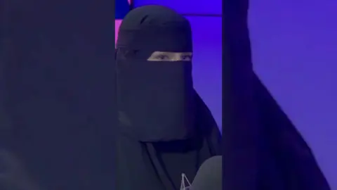 A woman wearing the niqab. She has a radio microphone in front of her.