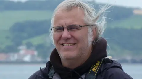 Robin Petch A smiling Robin Petch has long grey hair and wears a black jacket and  dark metal-framed spectacles. He looks windswept in front of a backdrop of the sea and green hills.