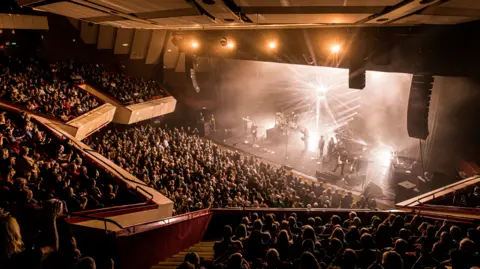 A general view picture of a band playing at the Hexagon with bright lights illuminated on the stage.