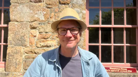 Tom Kennedy wears a straw hat, round glasses, and a light blue shirt. He stands in bright sunlight against a textured stone wall. Pink-trimmed windows frame the background, and a small red sign at the bottom right reads, “Hamptonne House As it appeared around 1736.”