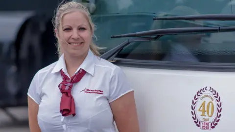 Alison in her white and red Grettons Coaches uniform, smiling and standing in front of a bus