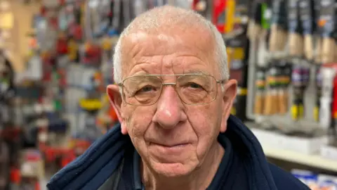 Elliot Deady/BBC A man - Neil Howells - looking straight at the camera. He is wearing glasses and a navy fleece and is standing in a hardware store with stock behind him.