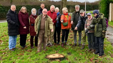 Meole residents standing on a grassy verge in front of a small oak stump, looking unhappy, with a sweet chestnut tree and hedgerow behind them. They wear jackets and some have headwear including caps and scarves. Many are pointing at the stump.